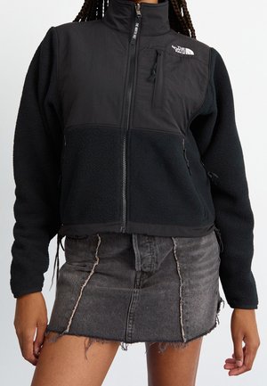 Persona que lleva una chaqueta de forro polar negra con cremallera y el logo de The North Face, y una minifalda vaquera negra con dobladillo deshilachado.