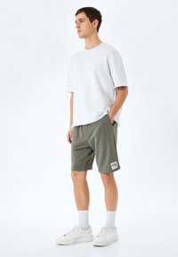 Ung mand står op, iført en løs hvid T-shirt med korte ærmer, olivengrønne shorts med en lap, hvide sokker og hvide sneakers foran en hvid baggrund.