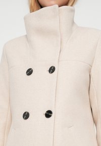Cappotto beige doppiopetto con colletto alto, realizzato in tessuto morbido, caratterizzato da bottoni neri e tasche laterali.