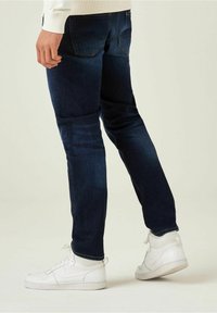 Man med mörkblå jeans och vita sneakers, står med ena foten något upphöjd mot en neutral bakgrund.