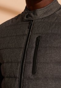 Lekka szara kurtka puchowa z wysokim kołnierzem, z poziomymi pikowanymi panelami i boczną kieszenią na zamek z czarnymi akcentami.