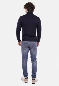 Maglione blu navy con colletto alto, texture lavorata a maglia e polsini a coste, abbinato a jeans azzurri e scarpe grigie.