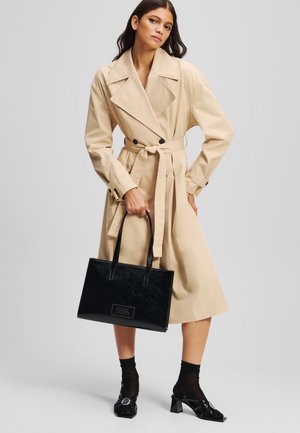 Cappotto beige con ampi risvolti, bottoni neri e vita cinturata, abbinato a una borsa a tracolla nera lucida con il logo. Scarpe con tacco nere e calze.