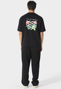Czarna koszulka z kwiecistymi grafikami i napisem "PAS DE MONACO" z tyłu, w połączeniu z czarnymi spodniami i jasnymi butami.