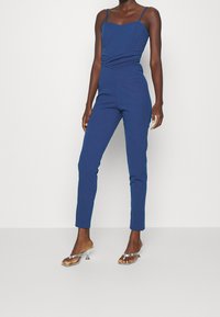 Kungslila jumpsuit med en åtsittande kropp, tunna axelband och rynkdetalj i midjan, matchad med metalliska klacksandaler.