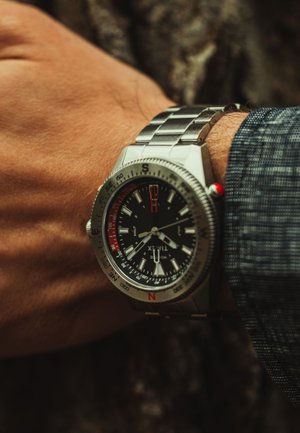 Montre en métal argenté avec un cadran noir, des index d'heure blancs, des accents rouges et une lunette à compas. Portée sur une main avec une surface texturée.