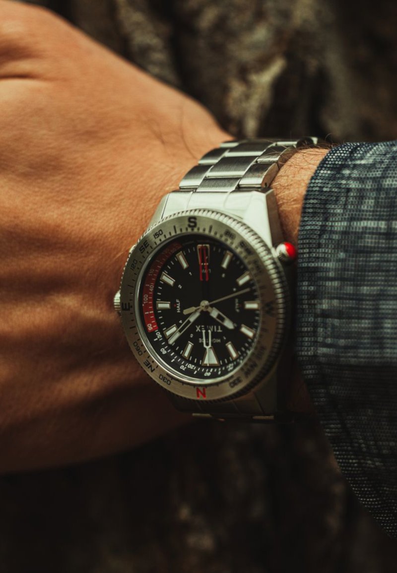 Orologio in metallo argentato con quadrante nero, indicatori delle ore bianchi, dettagli rossi e lunetta con bussola. Indossato su una mano con una superficie texturizzata.