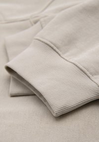 Sweat-shirt beige clair plié, montrant le poignet côtelé et la texture douce du tissu en détail rapproché.