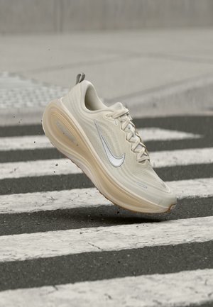 Beige Nike løbesko med hvid Swoosh-logo i luften over et fodgængerfelt på en asfaltvej.