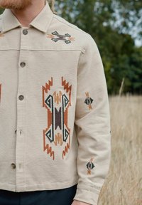 Beige geborduurde jas met geometrische patronen in bruin en groen, knoopsluiting en een standaardkraag met decoratieve accenten.