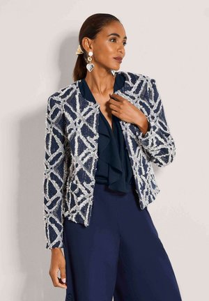 Vrouw draagt een getextureerd marineblauw en wit jasje met patroon over een marineblauwe blouse met ruches en bijpassende broek, met hartvormige oorbellen en paardenstaart.