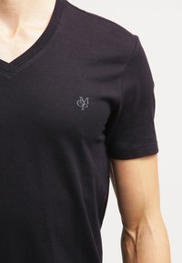 Hombre que lleva una camiseta negra de cuello en V y mangas cortas con un pequeño logo "MCP" en el pecho izquierdo frente a un fondo liso.