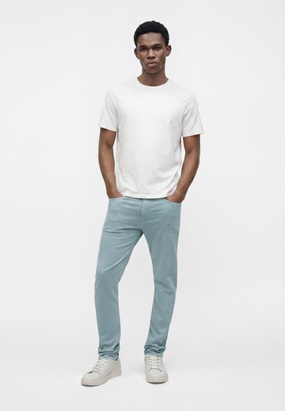 Jeune homme debout, les mains dans les poches, portant un t-shirt blanc uni, un pantalon bleu clair et des baskets blanches sur un fond blanc.