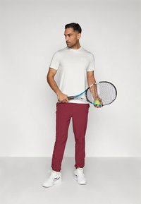 Muž v bílém tričku s krátkým rukávem, vínových kalhotách a bílých teniskách, držící tenisovou raketu a míček, se dívající na stranu.