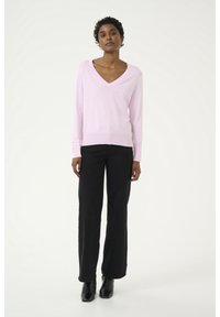Maglione rosa a scollo a V con maniche lunghe, caratterizzato da dettagli con bottoni sui polsini. Indossato con pantaloni larghi neri e stivaletti neri.