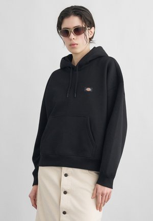 Persoon gekleed in een zwarte hoodie met voorzak en klein logo, beige rok met knopen en bruine zonnebril tegen een effen grijze achtergrond.