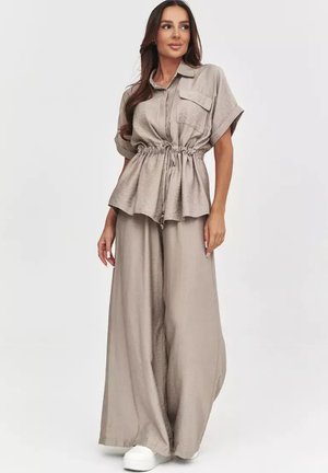 Kobieta w beżowej bluzce na krótki rękaw zapinanej na guziki z troczkiem w talii i szerokich spodniach, w połączeniu z białymi trampkami na jednolitym tle.