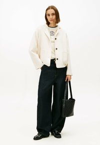 Roomkleurig, oversized jasje met grote knopen, gedragen over een licht topje; gecombineerd met een hooggesneden, donkere spijkerbroek en een zwarte gevlochten tas.