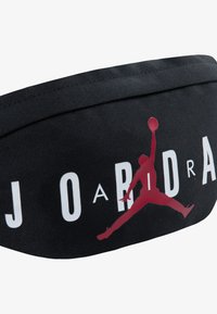 Uma faixa de cabeça de tecido preto com um grande logotipo vermelho Jumpman e um texto em branco em destaque a dizer "J O R D A N", com uma superfície texturizada.