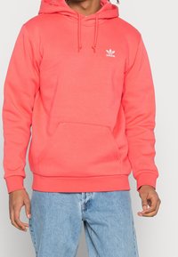 Korallrosa huvtröja med dragsko och kenguruficka; har en vit Adidas-logotyp. Materialet verkar mjukt, med ribbade ärmslut och nederkant.