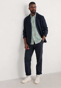 Blazer azul marino oscuro sobre una camisa con un patrón geométrico en verde y blanco, combinado con pantalones oscuros y zapatillas altas blancas. Ajuste y textura casual.