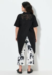 Zwarte blouse met korte mouwen met een doorschijnende, gemusterde rug, gecombineerd met wit tie-dye wijde pantalon en zwarte platte sandalen.