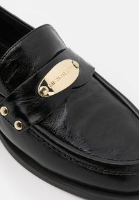 Mocassins en cuir noir présentant une plaque signature dorée bien visible et un matériel décoratif. Texture lisse avec des coutures le long de la semelle.