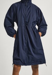 Tmavě modrý dlouhý kabát s předním zipem, vysokým límcem, elastickými manžetami a kapsami, nošený osobou, která má ruce v kapsách.