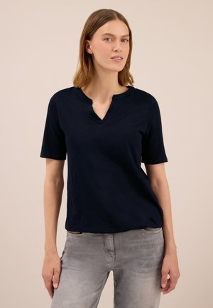Femme portant un t-shirt noir à manches courtes avec un petit col en V et un jean gris clair, se tenant devant un fond beige uni.