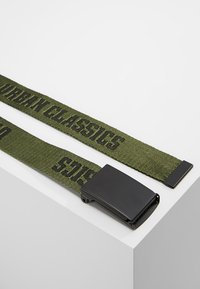 Zaļš austi josta ar melnu uzrakstu "URBAN CLASSICS", ar matētu melnu metāla sprādzi un taisnstūra galu.