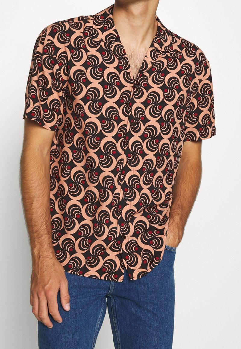Chemise à manches courtes présentant un motif tourbillonnant en noir et rouge sur un fond pêche. Confectionnée en tissu léger avec une coupe décontractée.