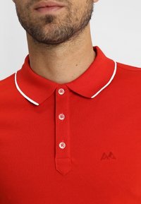 Röd pikétröja med strukturerad stickning, vit krage och fem knappar. Har en liten broderad logotyp på bröstet.