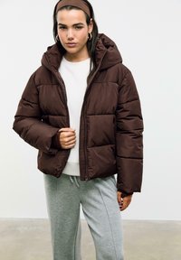 Smeđa puffer jakna s kapuljačom, s prepoznatljivim kvadratnim dizajnom i prednjim zatvaračem. Nosena preko bijele majice i sivi trenirke.