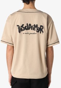 Camicia beige a maniche corte in rete con una vestibilità ampia. Presenta accenti neri e una scritta grafica sulla schiena che dice "La dichiarazione non è un prodotto."