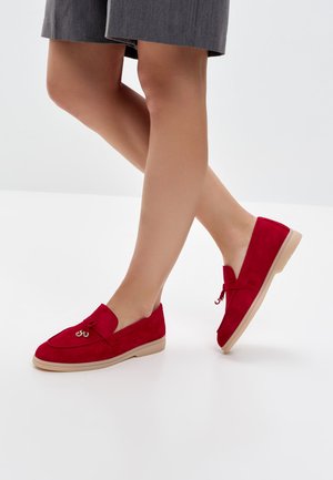 Piernas cruzadas con pantalones cortos grises y zapatos mocasines de ante rojo con suelas beige y pequeños borlas decorativas.