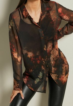 Femme portant une chemise transparente à manches longues avec des motifs abstraits automnaux sur un caraco noir et un pantalon en cuir noir.