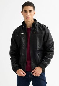 Chaqueta bomber de imitación cuero negra con cierre frontal, bolsillos en el pecho con cremallera, puños acanalados y hombreras, usada sobre una camisa burdeos.