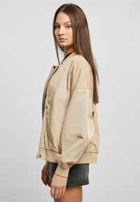 Giacca bomber beige con polsini e orlo a coste, dotata di zip frontale e chiusura con bottoni a pressione. Tessuto liscio e lucido; vestibilità rilassata.
