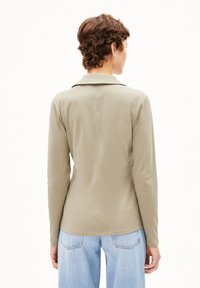 Camicia a maniche lunghe con colletto in morbido tessuto beige, dal tocco liscio, con una cucitura al centro e una vestibilità rilassata, abbinata a jeans azzurri chiari.