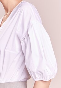 Blusa bianca a maniche a sbuffo con scollo a V profondo e tessuto testurizzato. Presenta una piccola tasca sul petto e dettagli arricciati alle maniche.