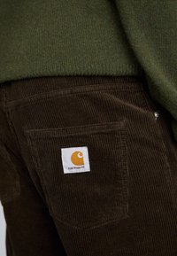 Hnedé kordové nohavice s textúrovaným povrchom, s bielou značkovou nášivkou a oranžovým logom na zadnej vrecku.
