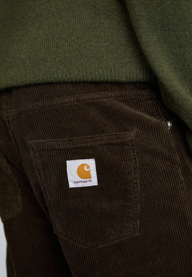 Braune Cordhose mit strukturiertem Finish, versehen mit einem weißen Markenetikett mit orangefarbenem Logo auf der Gesäßtasche.