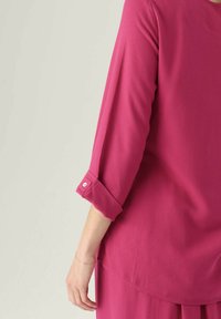 Blusa fucsia con maniche a tre quarti, caratterizzata da un colletto rotondo e un bottone decorativo sul polsino. Tessuto liscio e leggero.