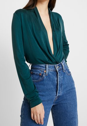 Mujer con una blusa de manga larga de color verde oscuro con un escote en V profundo, metida en unos vaqueros azules de talle alto.