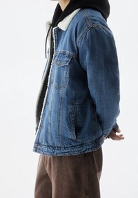 Persona con chaqueta de mezclilla azul con forro de vellón blanco sobre una sudadera con capucha negra y pantalones marrones, de pie con las manos detrás de la espalda.