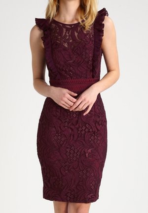 Femme portant une robe moulante sans manches en dentelle violet foncé avec des détails volantés aux épaules et un motif texturé.