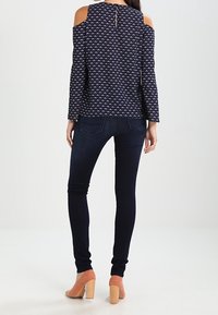 Navy cold-shoulder blouse with a geometric pattern, paired with dark skinny jeans and coral block heel shoes. Back view shows keyhole detail.