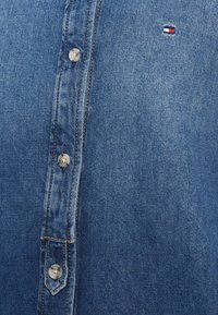 Πουκάμισο από τζιν σε ανοιχτό μπλε χρώμα με ασημένια κουμπιά, εμφανείς ραφές και μικρό λογότυπο σε κόκκινο, λευκό και μπλε στην επάνω αριστερή πλευρά.