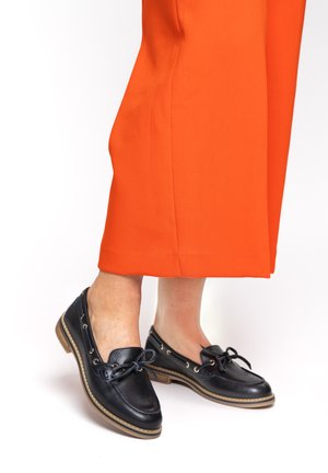 Woman wearing black leather boat shoes with light brown soles and orange cropped pants against a white background.