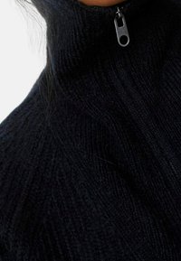 Zwart gebreide trui met rits rond de nek, voorzien van een geribbelde textuur en een metalen ritssluiting aan de hals. Close-upweergave benadrukt de stofdetails.